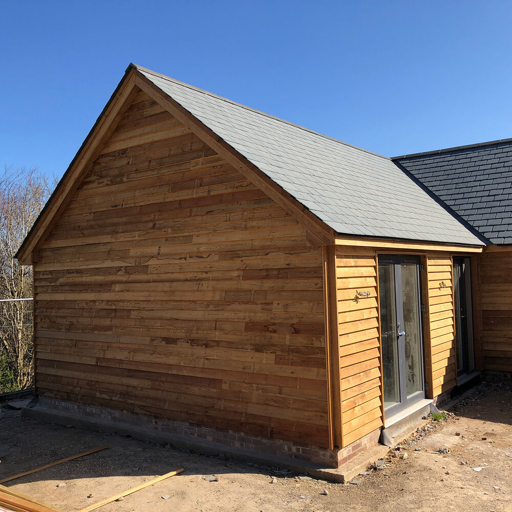 A newly built wooden shed with a pitched roof and large glass double doors, situated on a bare patch of ground under a clear blue sky.