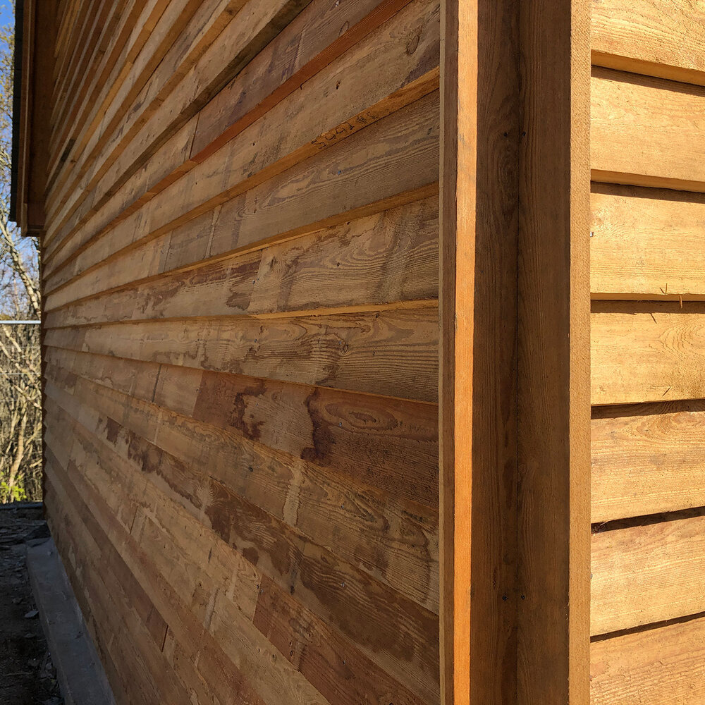 Close-up view of a wooden building exterior, showing horizontal wood siding with natural grain patterns and warm, sunlit tones. The image is taken from an angle emphasizing the texture and construction details.