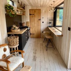 A cozy, modern tiny home kitchen with light wood walls and floors, black cabinets, open shelves, a small stove, and a window bar with two stools. A cowhide chair and wicker baskets add rustic charm.