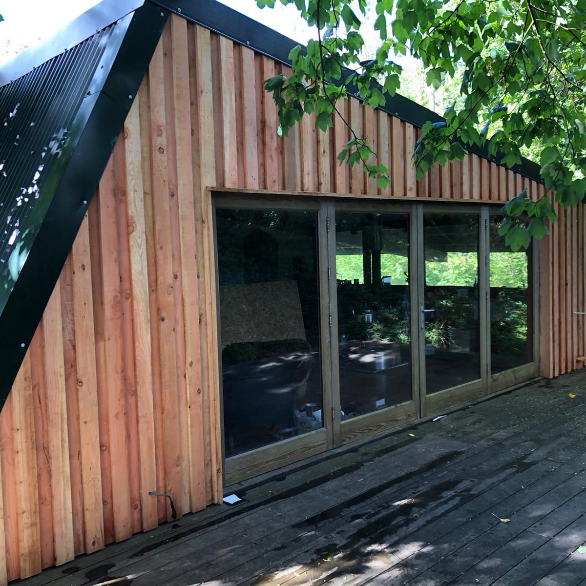 A small wooden cabin with vertical timber siding and large glass doors sits on a weathered wooden deck, shaded by leafy green tree branches.