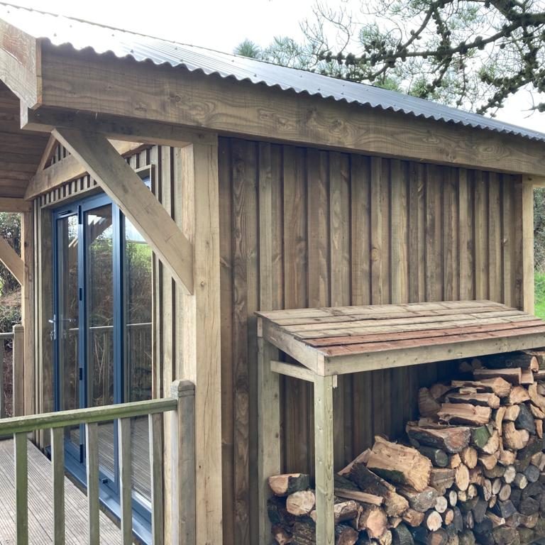 A wooden cabin with vertical paneling, glass doors, and a small covered area storing stacked firewood next to a railing. Trees and greenery are visible in the background.