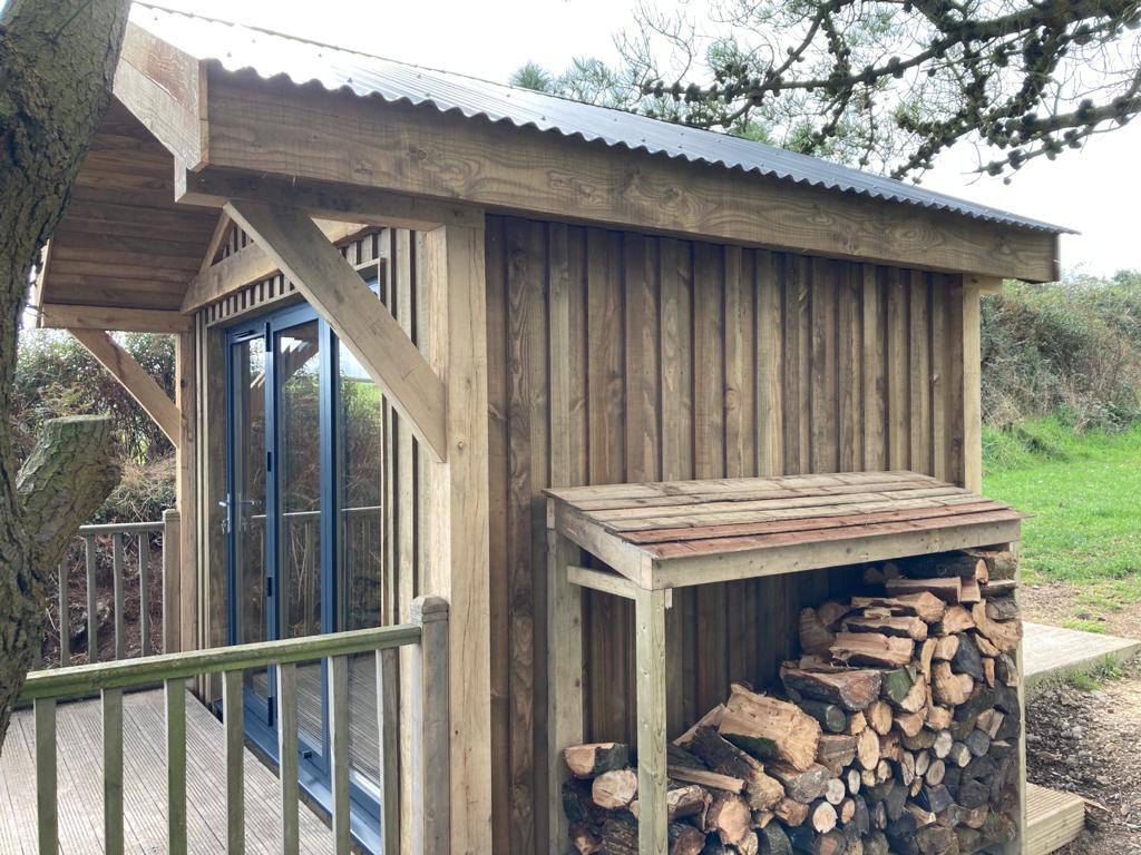 A small wooden cabin with a metal roof, double glass doors, and a stack of firewood under a shelter on a wooden deck, surrounded by greenery and trees.