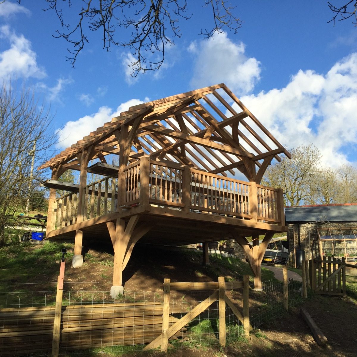 A wooden, open-framed gazebo is elevated on sturdy posts above the ground, with a partial roof structure and railings. It sits in a grassy area surrounded by trees under a blue sky with scattered clouds.