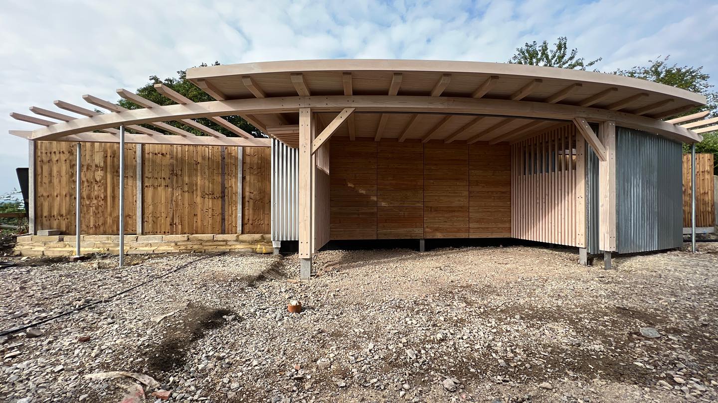 A modern wooden shelter with a curved roof and open sides stands on a gravel area, with a wooden fence and trees in the background under a partly cloudy sky.