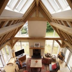A bright, spacious living room with high vaulted ceilings, exposed wooden beams, large windows, comfortable furniture, a dining table, and a fireplace, overlooking a green garden outside.